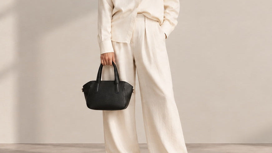 A person wearing a cream-colored linen two-piece outfit holds a small black leather handbag against a minimalist beige wall.