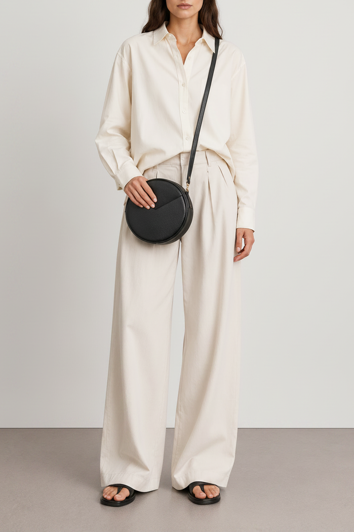 A model wearing a cream-colored long-sleeve button-down shirt and matching wide-leg trousers, styled with a black leather round crossbody bag and black slide sandals against a neutral studio background.