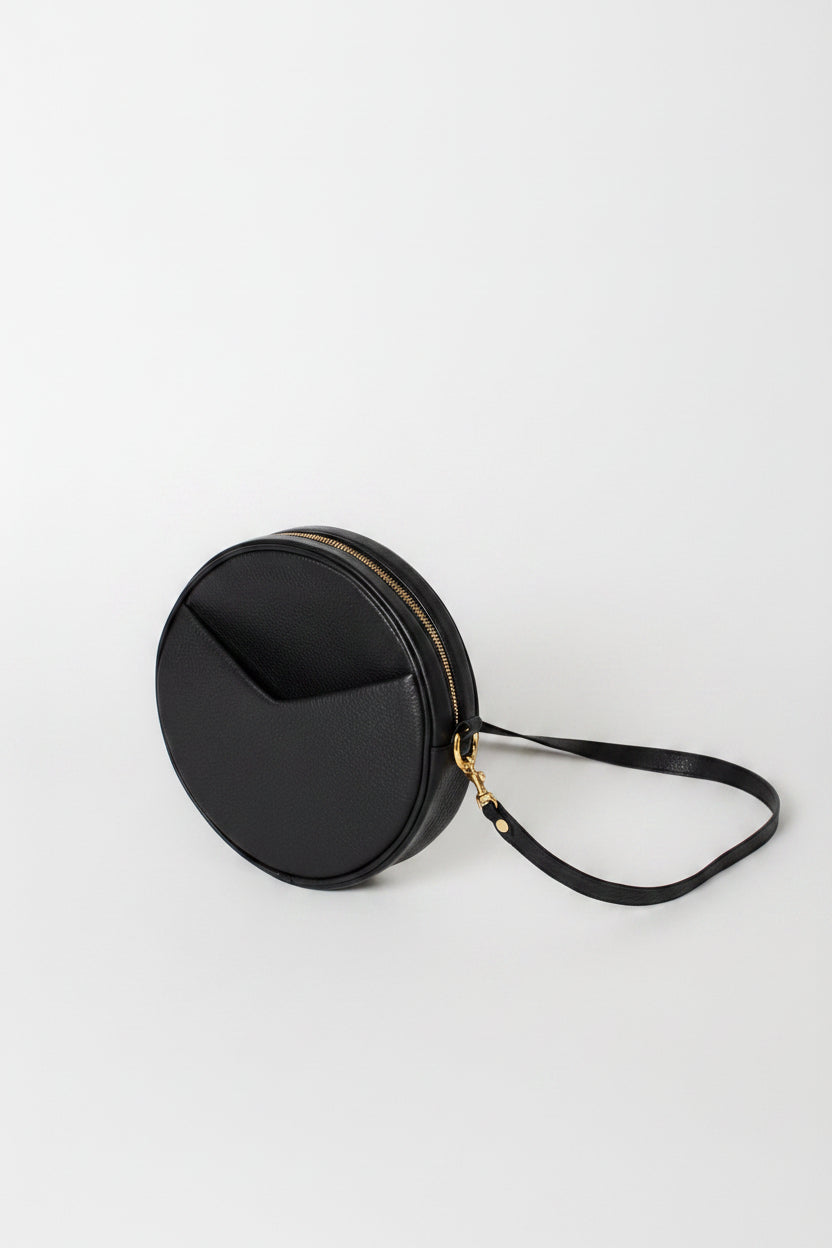 A black round leather crossbody bag with a gold-tone zipper and a thin black strap, photographed on a light gray studio background.