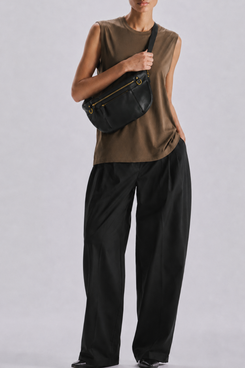 A model wearing a sleeveless brown top, black wide-leg pants, and a black leather commuter fanny crossbody bag with gold hardware, standing against a light gray studio background.