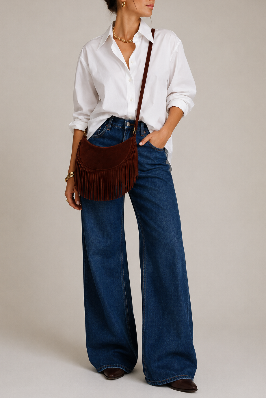 A model wearing a white button-down shirt, wide-leg blue jeans, and a dark brown suede fringe crossbody bag.