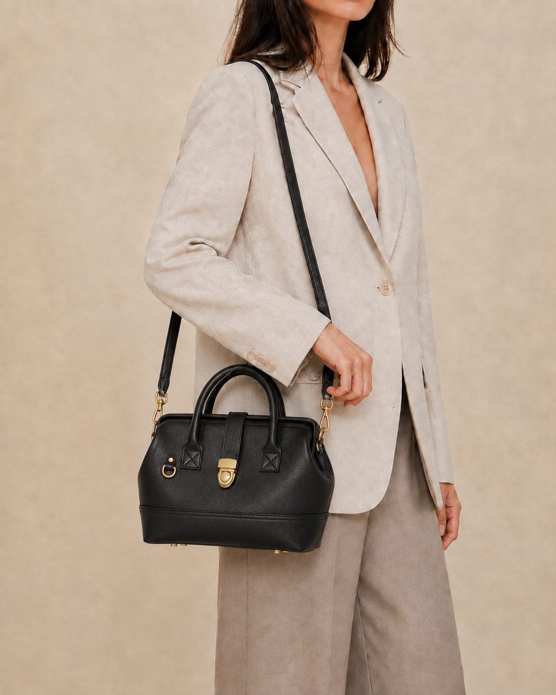 A woman wearing a light beige blazer and matching trousers carries a structured black leather Doctor Bag with gold-tone hardware and a black shoulder strap. 