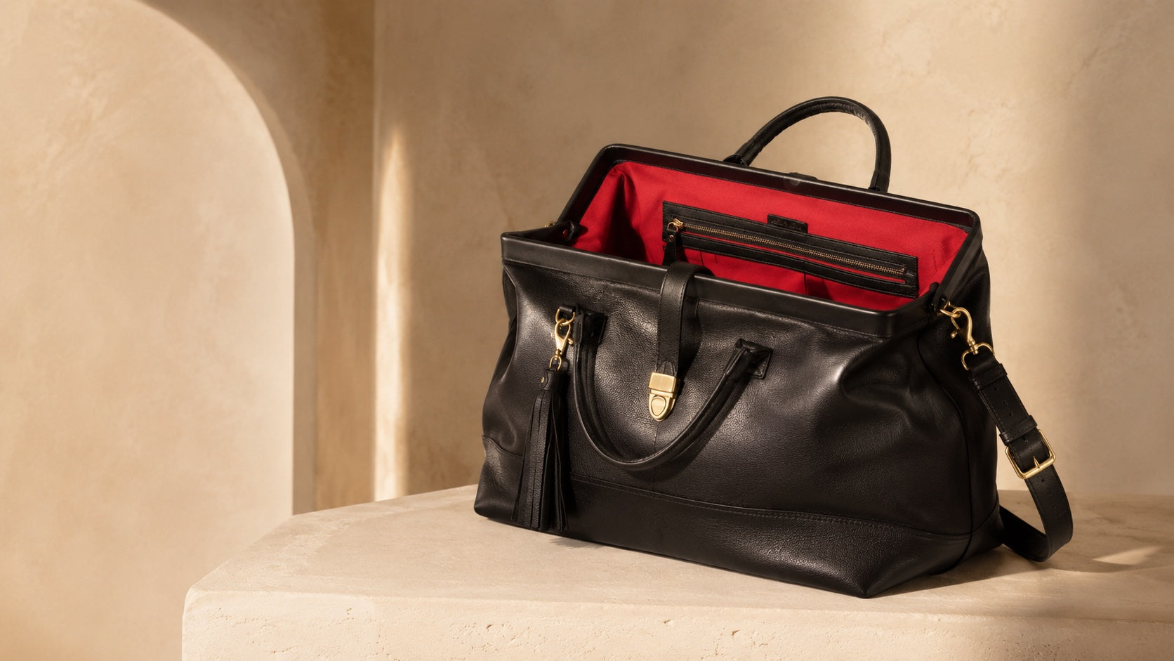 A black leather Doctor weekender duffel bag with a red interior lining, displayed open on a beige stone pedestal in warm sunlight.