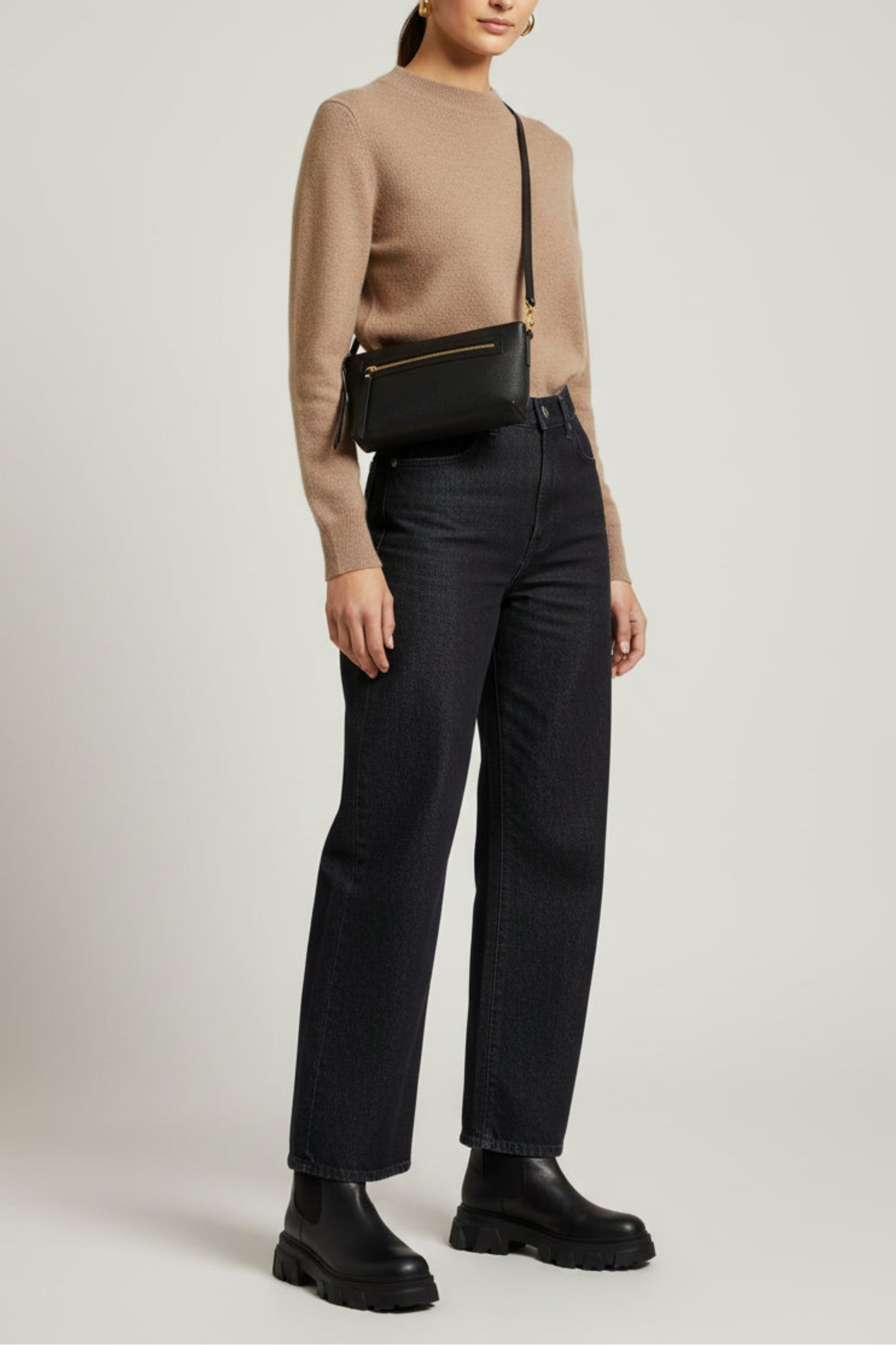 Woman wearing a beige sweater, black pants, and black shoes with a black handbag on a light gray background