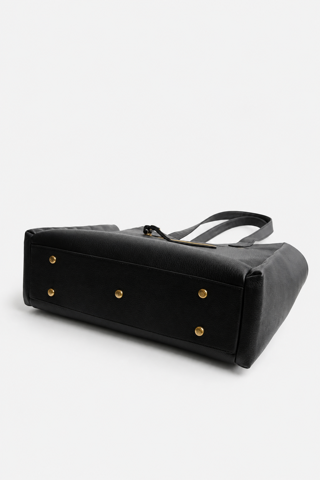 A black leather tote bag with gold-tone rivets on the bottom base and a matching black shoulder strap, photographed on a clean white studio background.