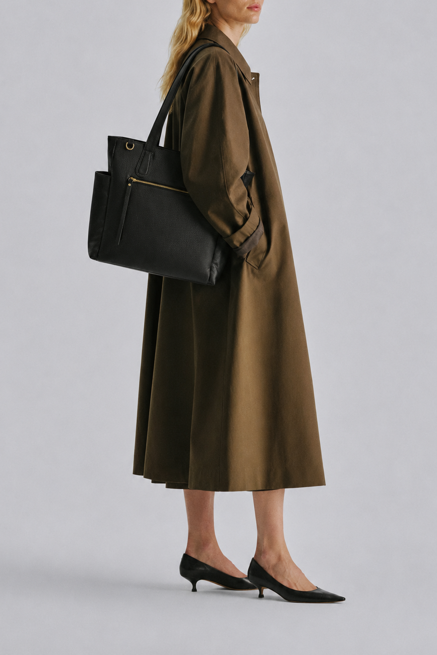 A woman in a long olive-brown coat and black pointed-toe heels carries a large black tote bag over her shoulder, photographed against a plain light gray studio background.