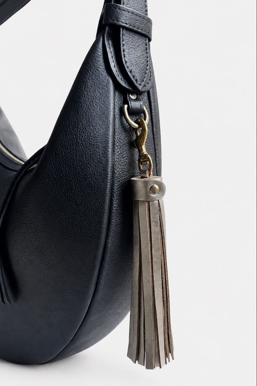 A black pebbled leather crescent-shaped shoulder bag with a matching black strap, gold-tone hardware, and a gold leather tassel charm, shown against a light gray background.