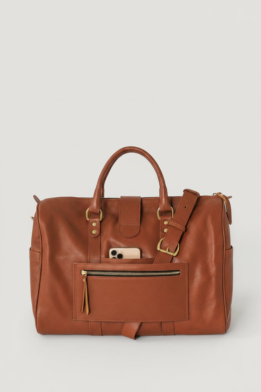 A brown leather duffel bag with gold-tone hardware, featuring dual top handles, a front zippered pocket, and a shoulder strap, with a pass-through luggage pocket. 