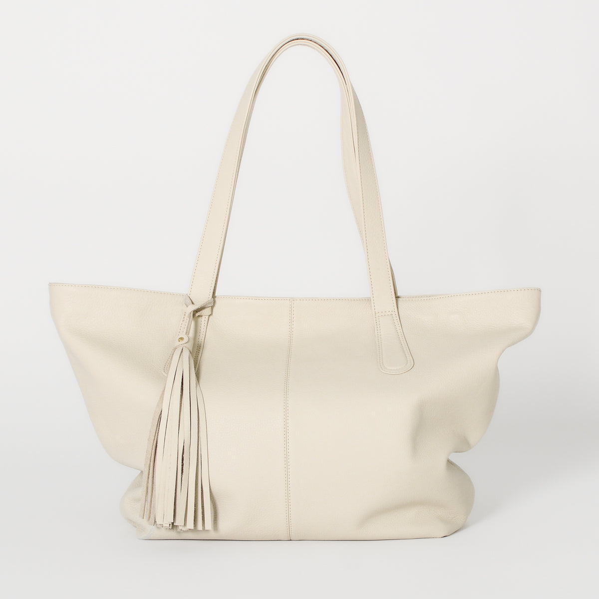 A off-white leather tote bag with solid brass hardware, a secure zipper closure, and a large detachable tassel.