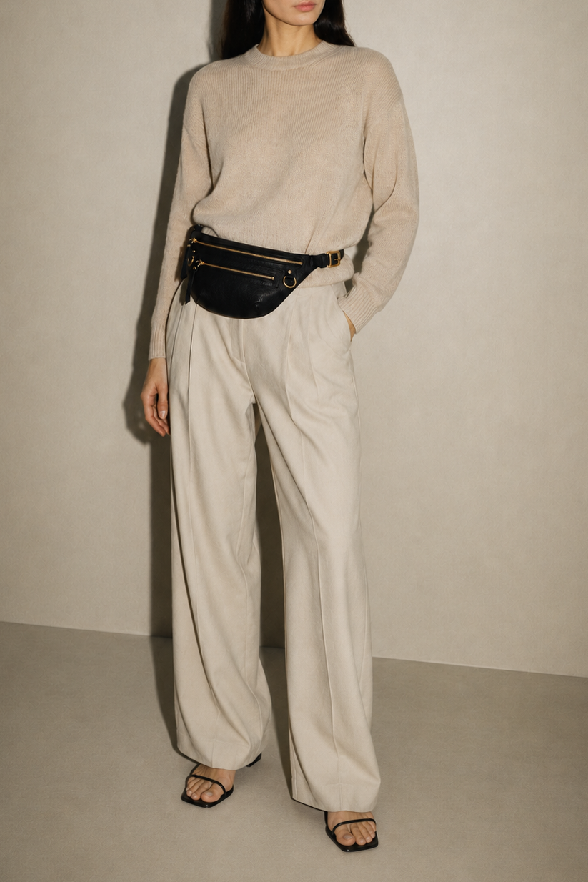 Nomad Fanny Pack in black full-grain leather worn at the waist on a full-length model styled in a cream knit sweater and wide-leg trousers, showing the bag's slim profile and scale as a hands-free belt bag for everyday wear.