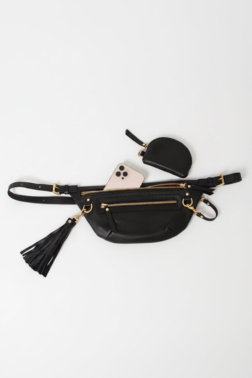 Nomad Fanny Pack in black leather laid flat with phone tucked in main compartment, styled with a leather tassel and small coin pouch, showcasing storage capacity and accessory compatibility for everyday essentials.
