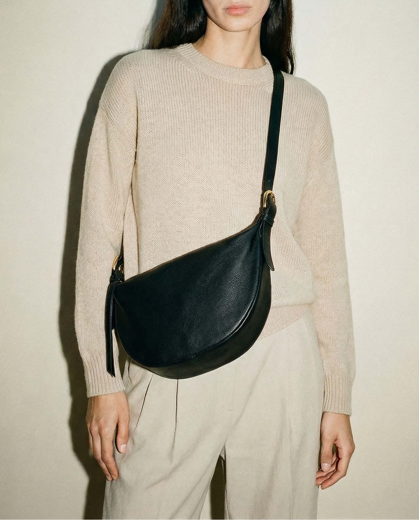 Woman wearing a beige sweater and light pants with a black shoulder bag against a plain background