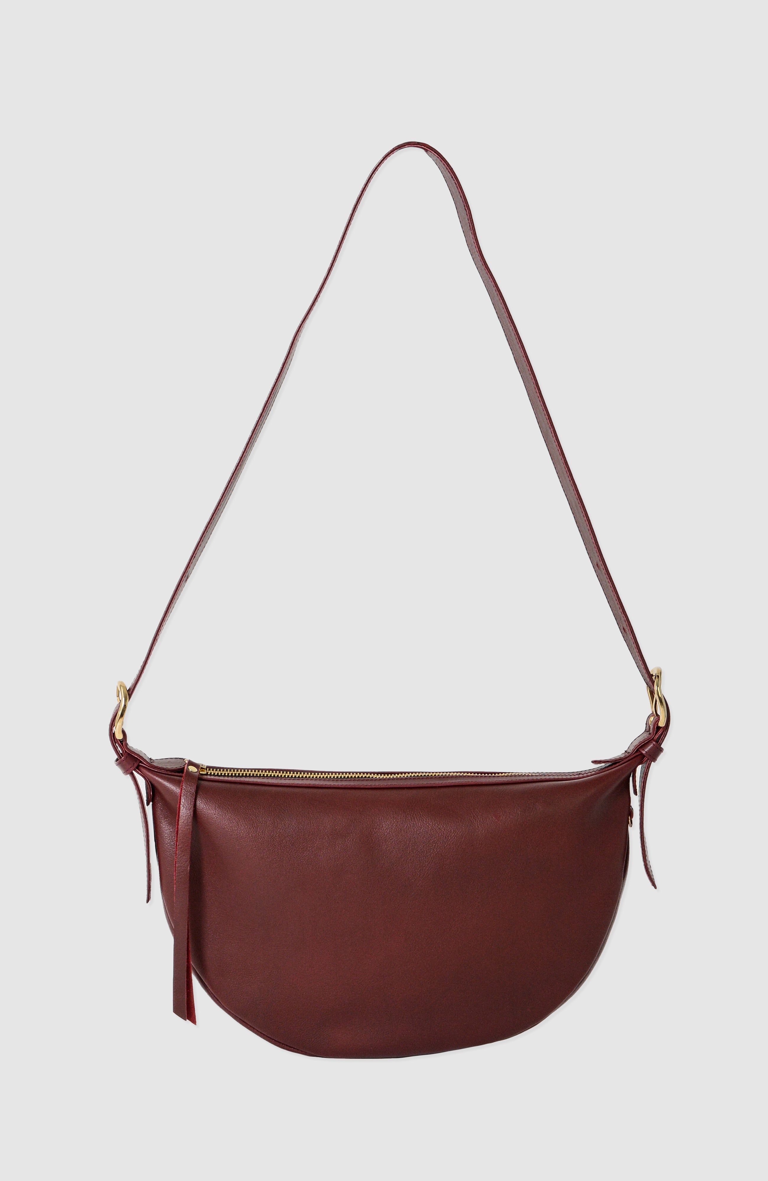 Nova Sling Bag in full-grain leather with brass hardware, softly structured curved silhouette, zipper closure, interior compartments, and adjustable strap.