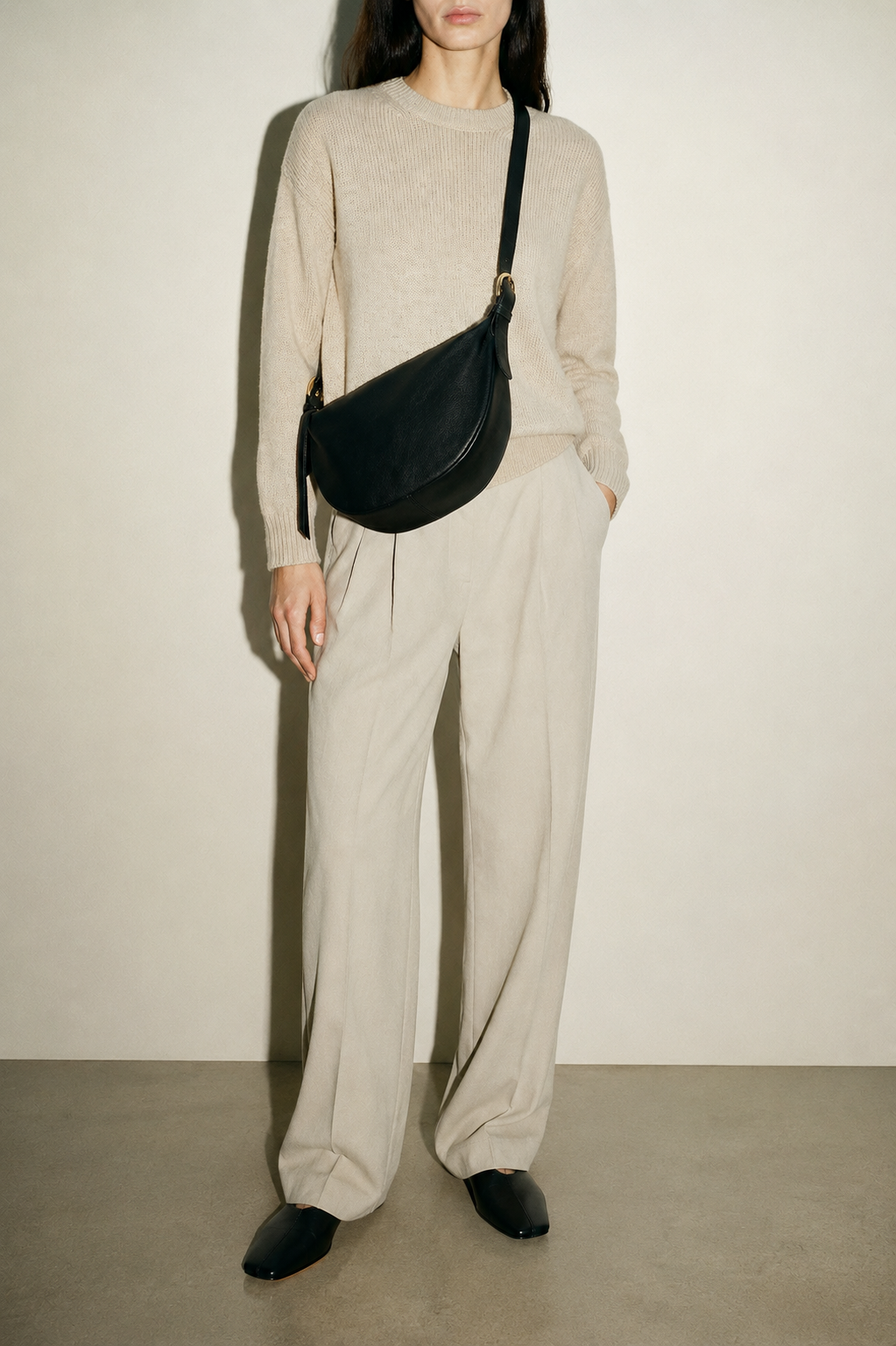 A model wearing a beige knit sweater and matching wide-leg trousers, styled with a black leather crescent-shaped crossbody bag and black pointed shoes, standing against a plain beige wall. The bag in this image is the TAH Nova Sling Bag.