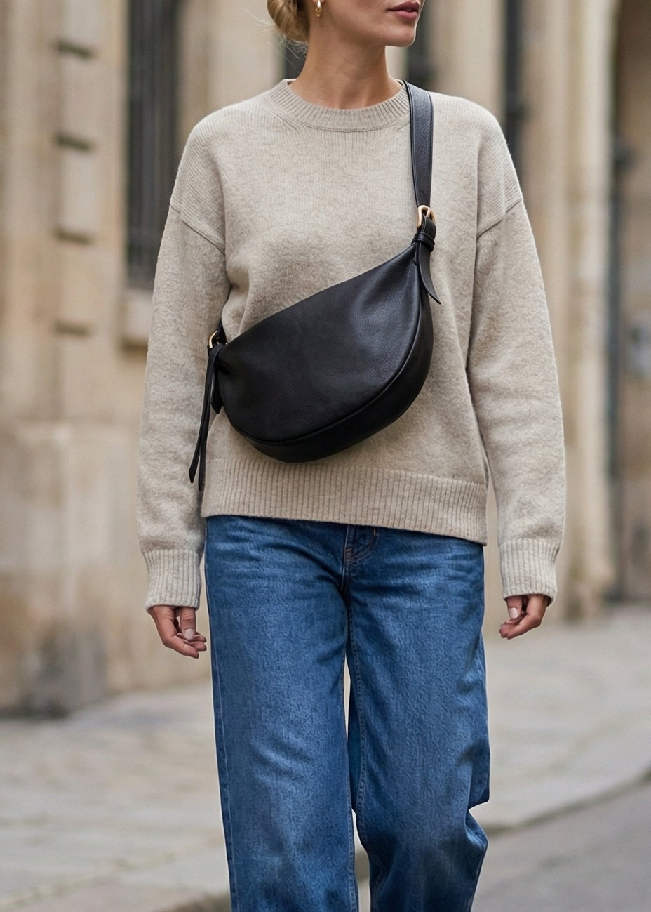 Woman walking on a street wearing a beige sweater, blue jeans, and carrying a black bag.
