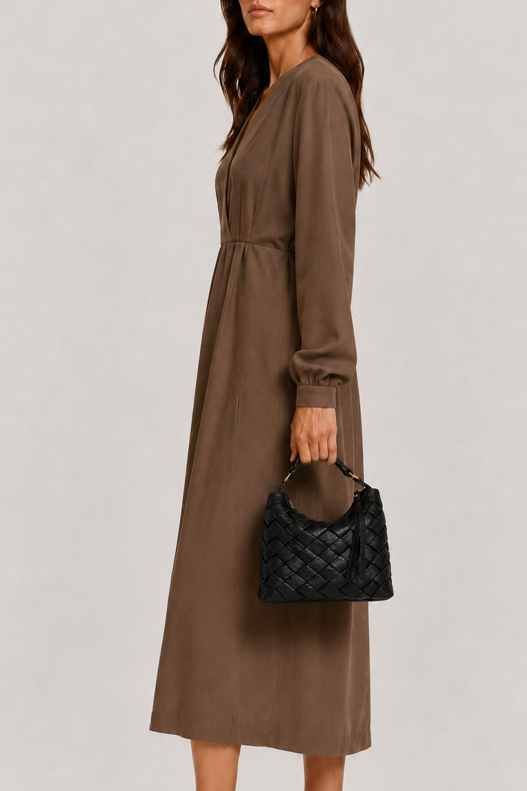 A woman modeling a long-sleeve brown midi dress with a gathered waist, paired with black pointed flats and a black quilted handbag, set against a plain light gray studio background.