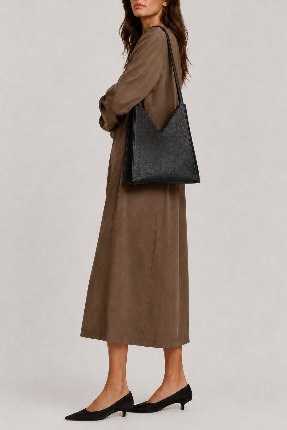 A woman wearing a long brown dress, black sunglasses, and black pointed-toe heels carries a large black leather shoulder bag against a plain light gray studio background. The bag is the Vie Tote bag by TAH Handbags.