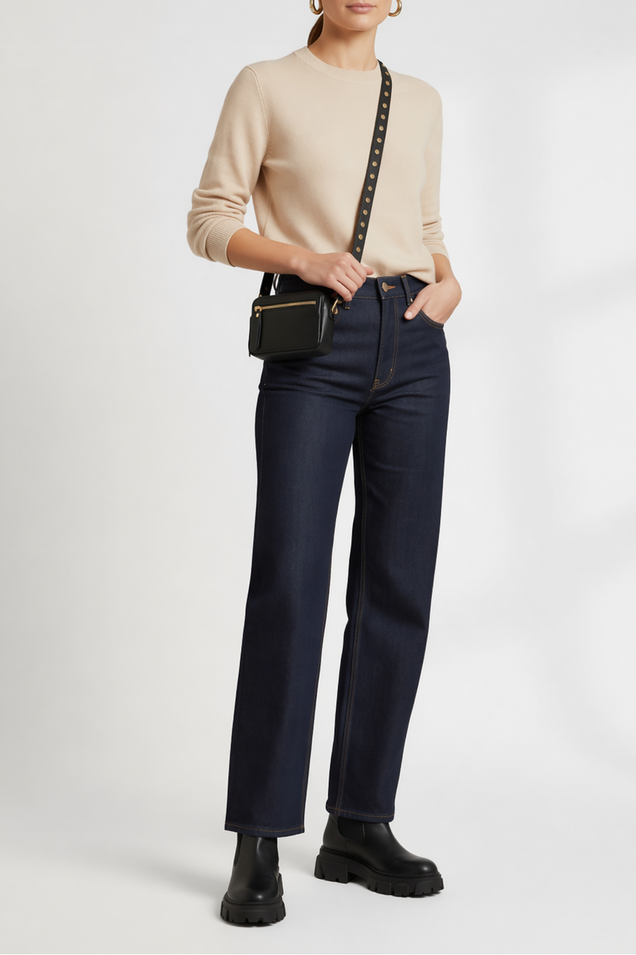 Model wearing the black leather compact Crossbody bag with a gold studded strap, and exterior zipper, styled with a neutral sweater, dark denim, and black boots.