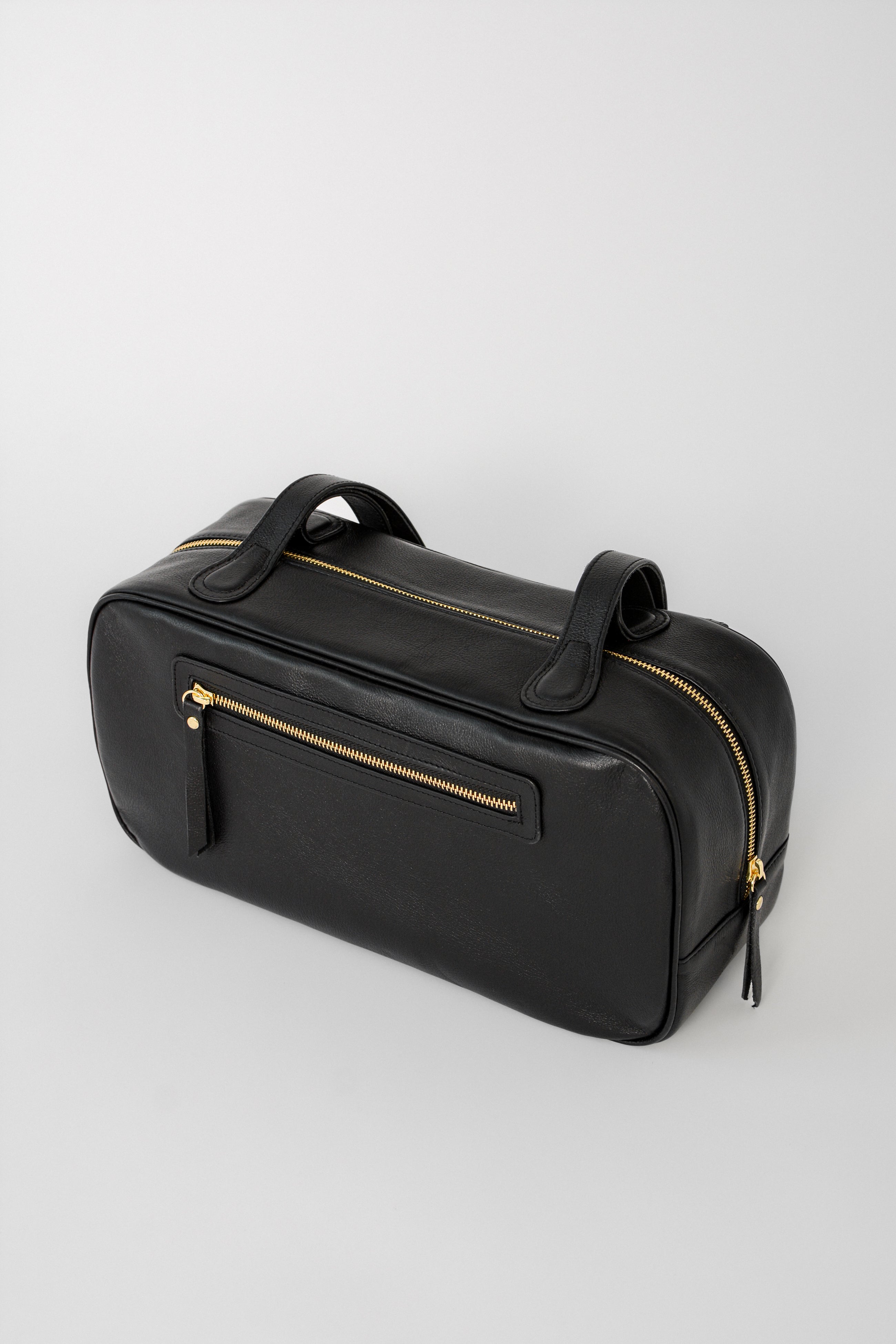 Black leather shoulder bag with a structured rectangular silhouette, shown at an angled side view with a top zip closure, exterior zip pocket, and long straps, finished with a minimalist, refined design.