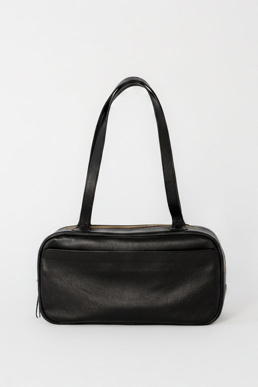 Black leather bowler shoulder bag with a structured rectangular silhouette, long straps, and top zip closure, designed as a minimalist everyday bag for work, travel, and daily use.