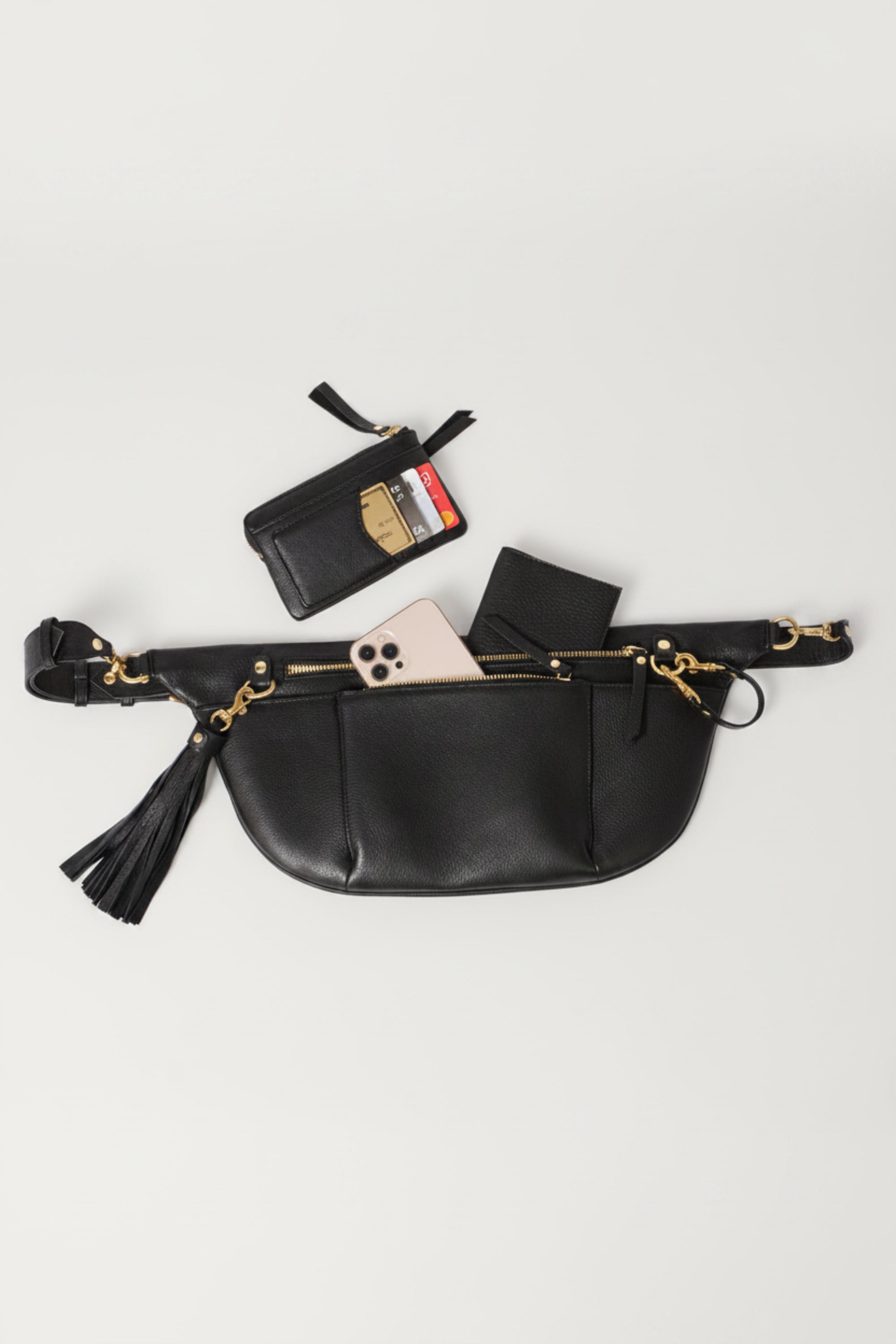 Black leather commuter sling bag with zip compartments and gold hardware, shown with phone, wallet, and card holder to highlight organized storage for everyday essentials and travel.