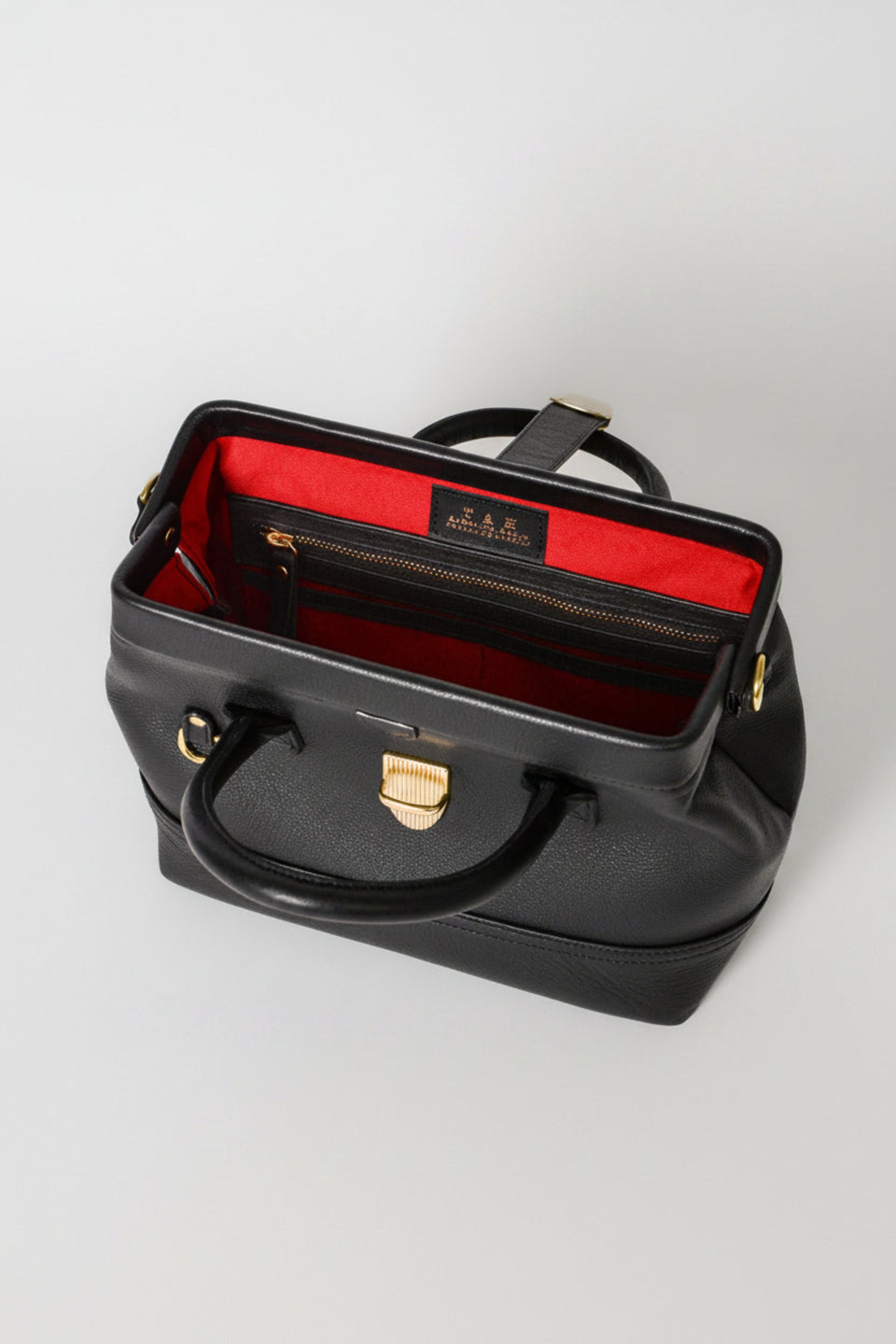 Interior view of the Doctor Bag in black leather, showing a structured metal opening with red lining and a zip pocket