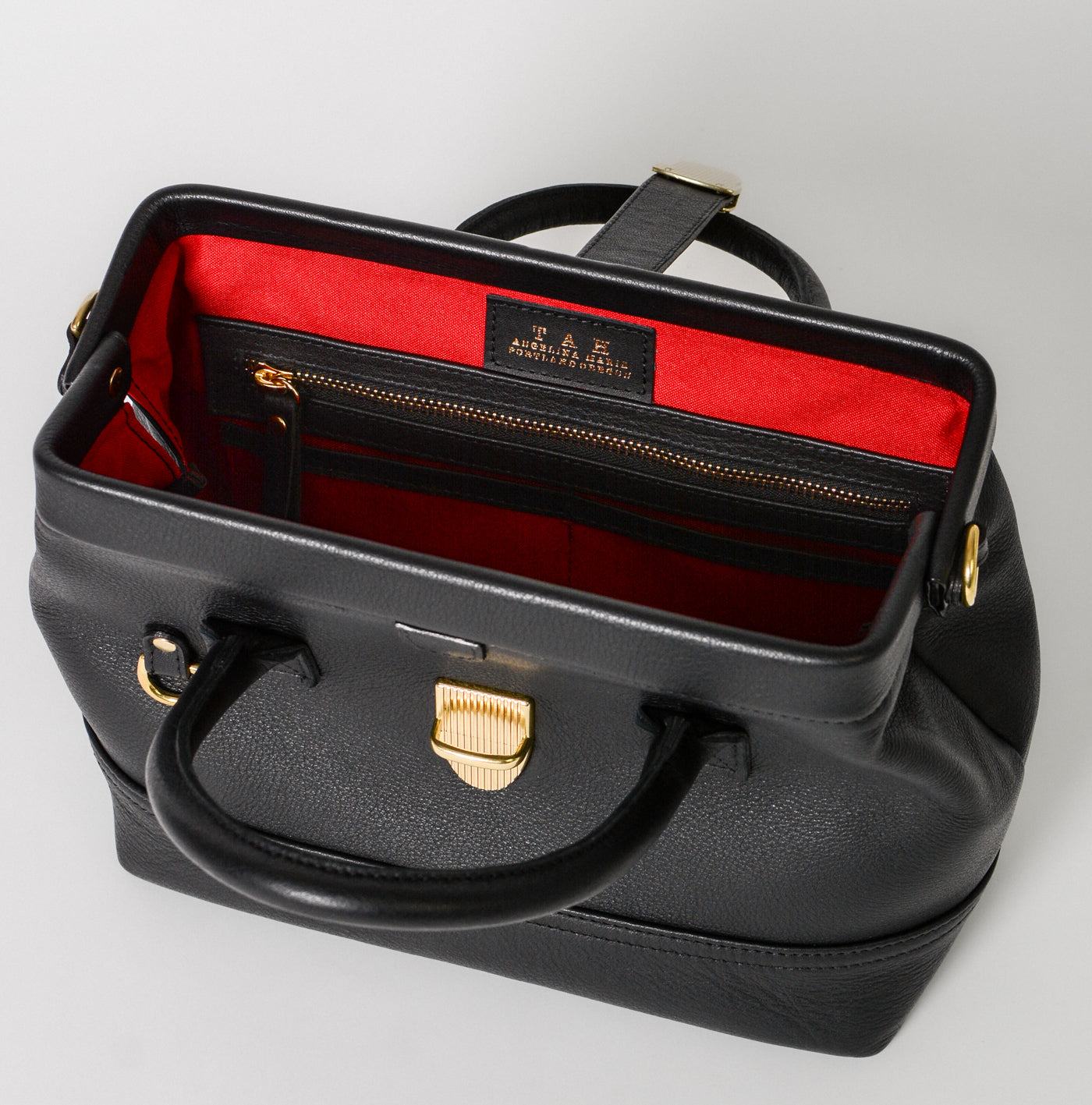 Interior view of the Doctor Bag Petite in black leather, showing a structured opening with red lining, a zip pocket, and branded leather tag.